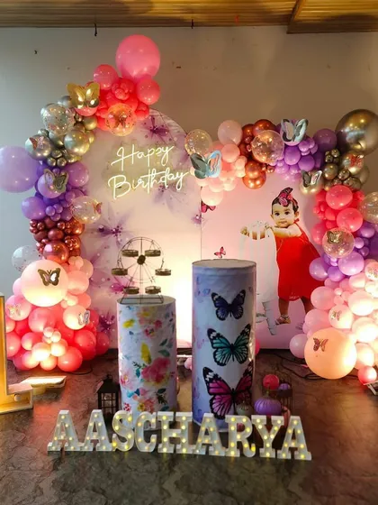 This elaborate butterfly theme for a first birthday uses a mix of pink, purple, and chrome balloons. The setup includes a personalized backdrop, themed plinths, and large light-up letters for the name and number '1'.