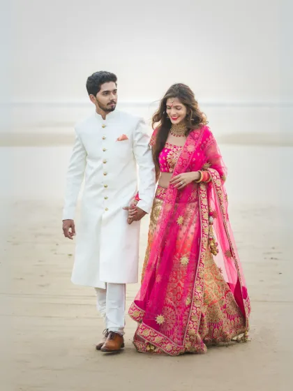 An elegant beach pre-wedding shot, with the couple dressed in coordinated white and pink traditional outfits, walking together by the sea.