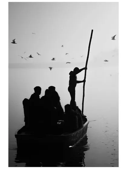 A black and white silhouette of a boatman steering his vessel through the foggy morning at Yamuna Ghat, surrounded by birds.