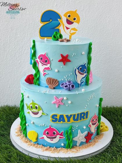 Doo doo doo! This two-tiered Baby Shark cake is an underwater adventure. It's decorated with the whole shark family using a mix of edible prints and fondant details, set against a blue whipped cream ocean.