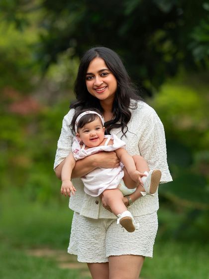 A mother in a lovely white outfit holds her smiling baby. The natural green background makes their light-colored clothes pop.