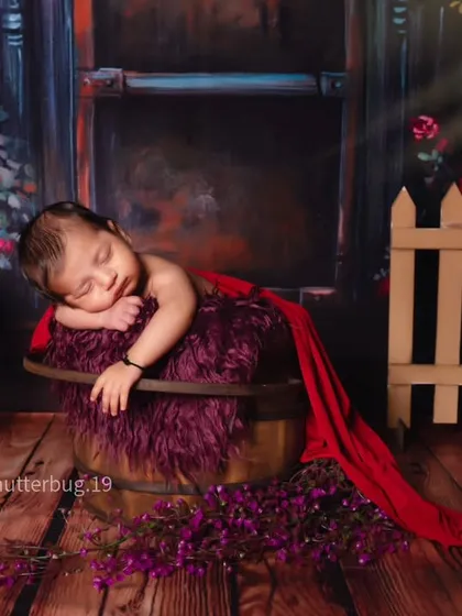 A side view of the dark floral setup, showing the baby in a peaceful sleeping pose with a dramatic red cloth draping from the bucket.
