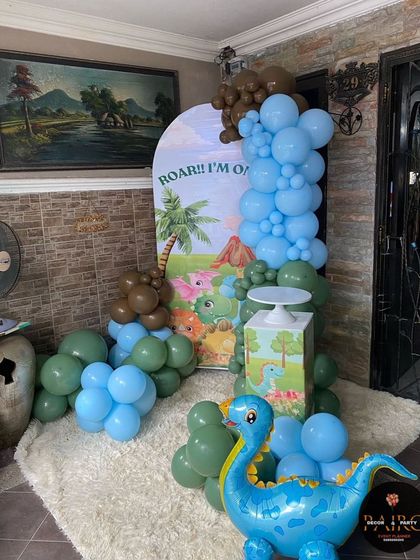 A cute "Roar! I'm One" dinosaur-themed birthday setup. This decoration uses an arched backdrop with a prehistoric scene, green and blue balloons, and a dinosaur foil balloon for a playful touch.