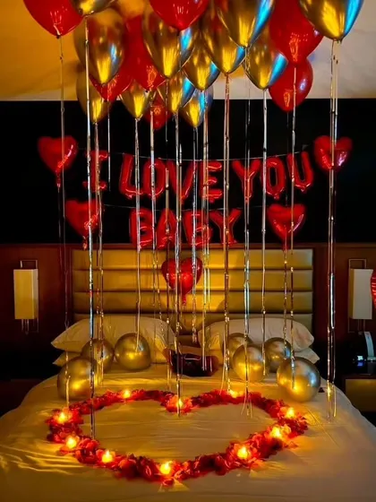 A classic romantic setup for an anniversary or birthday. The decor includes red and gold balloons, "I LOVE YOU BABY" foil letters, and a heart made of petals and candles on the bed.