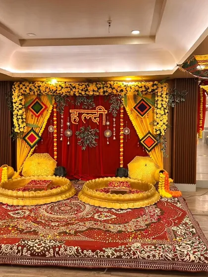 A Haldi setup in a banquet hall, featuring a red and yellow backdrop, traditional umbrellas, and two golden bowls for the ceremony.