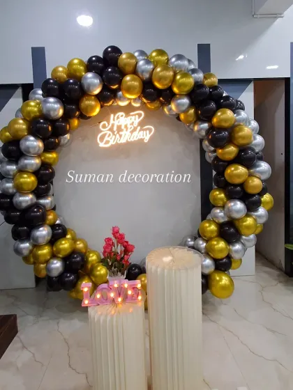 A chic and modern birthday setup using a circular balloon arch in black, gold, and silver. The decor is completed with a neon sign and pedestals for the cake.