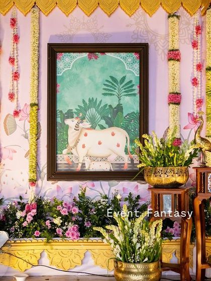 Another detail from the naming ceremony decor, showing a framed piece of traditional Indian art set against the larger painted backdrop, surrounded by fresh flowers and banana leaves.