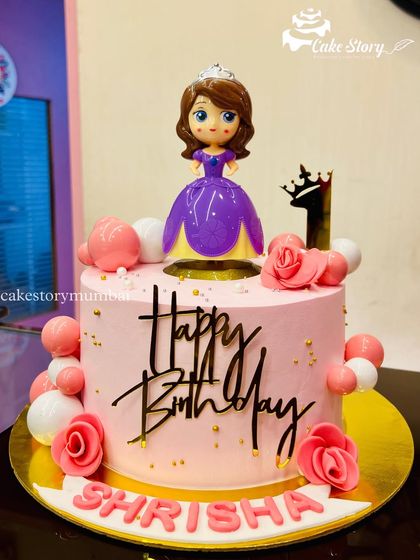 A pretty pink cake for Shrisha's first birthday, featuring a Princess Sofia topper. The design is decorated with pink and white edible pearls and a stylish 'Happy Birthday' script.