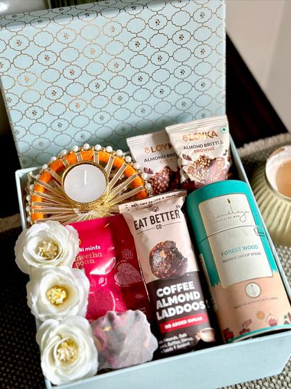 This elegant blue Moroccan-print box contains a lovely assortment of festive items. It features a decorated tealight holder, almond brittle, flavored chocolate ladoos, and fragrant dhoop sticks for a complete sensory experience.