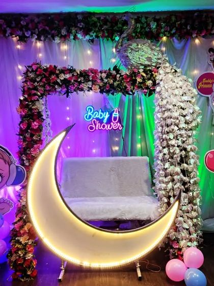 A modern baby shower setup with a lighted moon prop in front of a traditional decorated swing. The 'Baby Shower' neon sign and character cutouts complete the look.