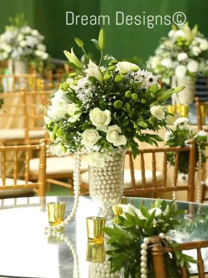 A beautiful mirrored tabletop reflecting a classic centerpiece. The arrangement of white lilies and roses is set in a pearl-covered vase, with pearl strings draping from the chairs to complete the elegant, pearlescent theme.