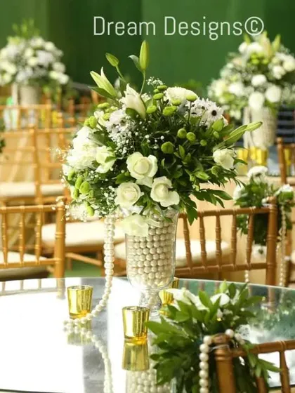 An elegant table setup featuring a mirrored tabletop that reflects the beautiful centerpiece. The vase is adorned with pearls, and the chairs are decorated with pearl strings for a touch of classic glamour.