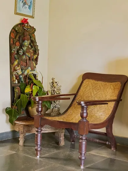 A classic, colonial-style planter's chair paired with a traditional carved wooden sculpture. This corner vignette is rich with history and character, reflecting the heritage of the Coorg region.