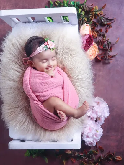 A unique pose showing a smiling baby girl comfortably suspended in a pink sling from a miniature bed prop, surrounded by flowers.