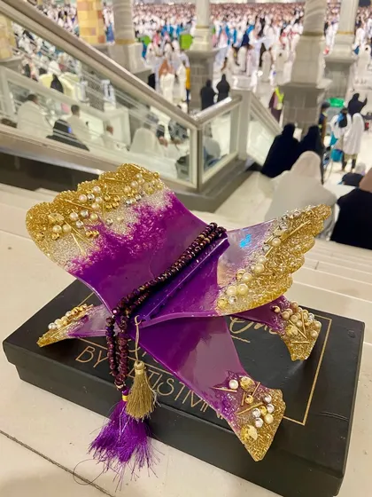 This striking Quran holder is made with a vibrant purple and gold resin design. It comes with a matching crystal tasbeeh and is shown here at the holy city of Makkah, adding to its spiritual significance.