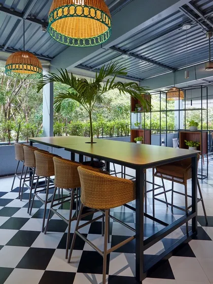 Biophilia integrated into a corporate cafe. This covered terrace features a checkerboard floor, rattan furniture, and a large palm tree, creating a relaxed, resort-like atmosphere.