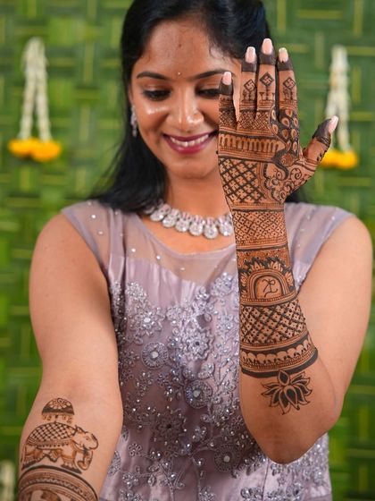 A happy client showing off her bridal mehendi, which includes a personalized initial and an elephant on the upper arm.