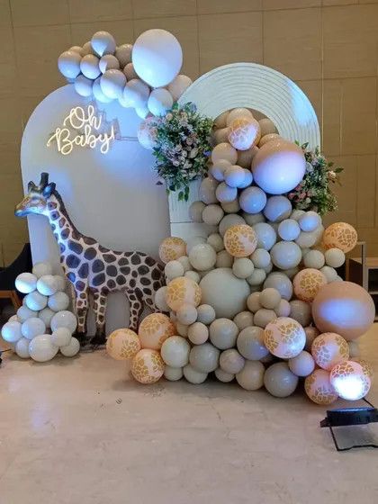 A chic safari-themed baby shower with an 'Oh Baby' neon sign. The decoration uses muted green and beige balloons with leopard print accents and a large giraffe prop for a stylish jungle vibe.