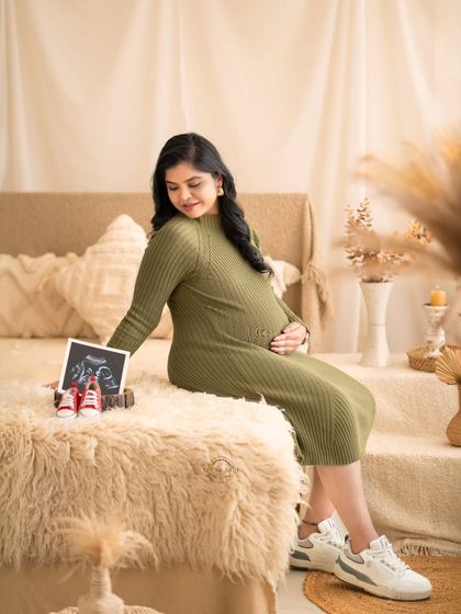 A cozy and sweet portrait of a mom-to-be in an olive green knit dress, looking down at her sonogram picture and a pair of baby shoes.