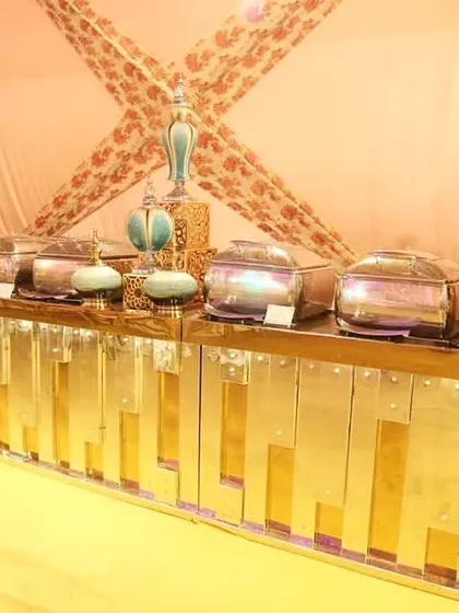 A glamorous buffet counter with a metallic gold finish and intricate details. The setup includes silver chafing dishes and decorative golden goblets under a draped pink canopy.