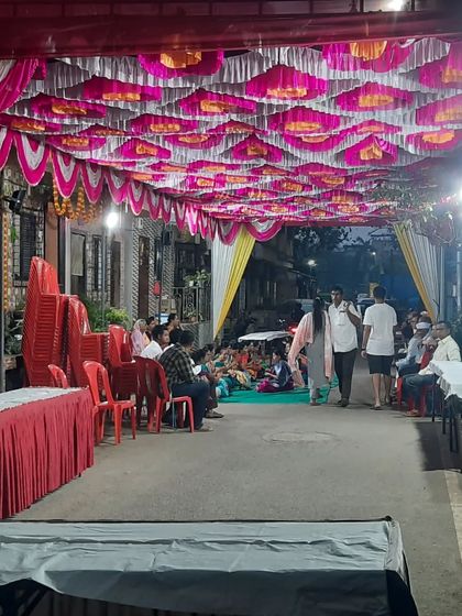 An outdoor event space with a decorated pandal ceiling. Guests are seated on the floor and on chairs, showing a flexible arrangement.