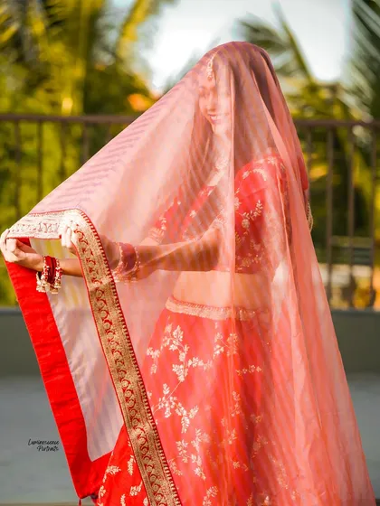 The bride playfully holds her sheer dupatta, creating a beautiful silhouette against the bright outdoor light.