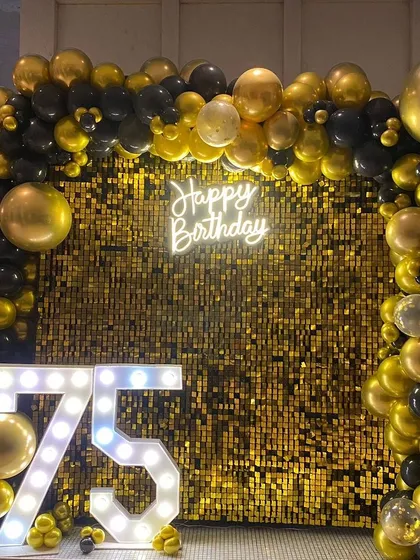 Celebrate milestone birthdays in style. This 75th birthday party features a glamorous gold shimmer wall, a black and gold balloon arch, a 'Happy Birthday' neon sign, and large light-up marquee numbers.