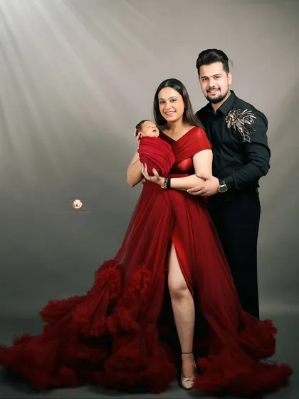 A glamorous family portrait celebrating a new beginning. The parents smile warmly while holding their newborn, with the mother's stunning red gown creating a bold and beautiful statement in the studio.