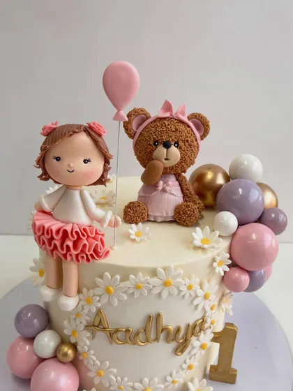 A soft pastel dream for a first birthday, featuring a hand-sculpted doll and a textured teddy bear, surrounded by a cascade of pastel and gold spheres and a wreath of daisies.