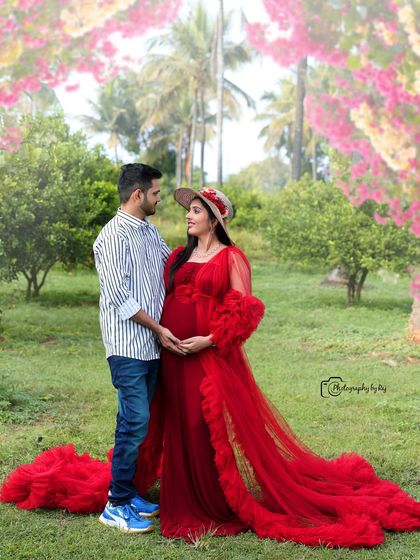 A romantic couple's portrait in a garden setting. The mother-to-be is wearing a red ruffled gown and hat, creating a classic and elegant look.