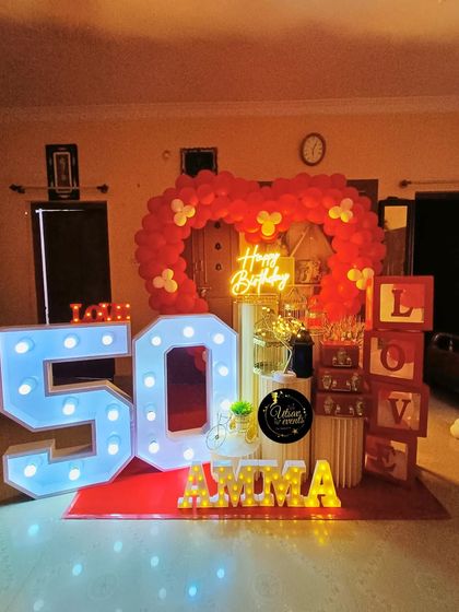 A surprise 50th birthday party for 'Amma' with a red balloon arch and large light-up marquee numbers. The 'LOVE' blocks add a heartfelt touch.
