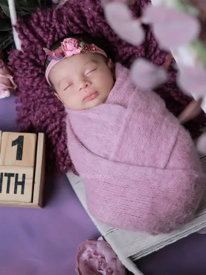 A one-month-old baby sleeps peacefully, swaddled in a purple wrap. Our newborn sessions are designed to keep your baby comfortable and secure while we capture these quiet moments.