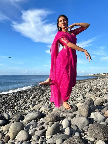 A dance pose in a bright pink saree on a pebble beach. The contrast between the vibrant color of the saree and the blue sky creates a visually striking image for fashion and travel campaigns.