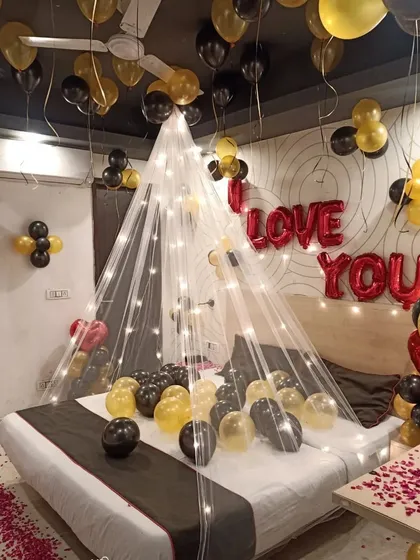 A romantic room setup with a fairy light canopy over the bed. 'I LOVE YOU' foil letters on the wall and scattered balloons in black and yellow create a sweet and intimate surprise.