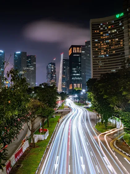 A long-exposure shot of a Singapore highway at night, transforming vehicle headlights and taillights into brilliant white streaks of light against the modern cityscape.