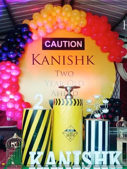 A creative construction theme for a second birthday party. The setup includes a bold "CAUTION" sign on a yellow backdrop, balloon arches in red, yellow, and black, and custom props like themed barrels and vehicle cutouts.