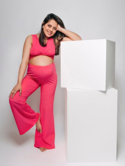 A fun and vibrant studio portrait of a mom-to-be in a bright pink two-piece outfit. Her joyful smile and relaxed pose against the clean white blocks create a modern and playful look.