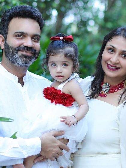 A classic family portrait taken outdoors, with a mother and father holding their daughter against a lush green background.