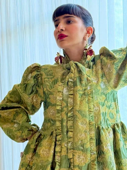A close-up of the green brocade dress, highlighting the ruffled details and statement earrings.