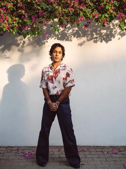 A full-length portrait of Zahan Kapoor standing under a bougainvillea tree. The shadows and flowers create a beautiful, natural frame.