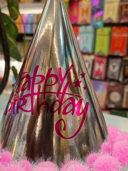 A metallic silver birthday hat with pink 'Happy Birthday' text and a cute pink pom-pom trim. It's a stylish and fun party hat that adds a touch of sparkle to the celebration.