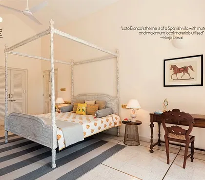 A bedroom in the Loto Bianco villa, designed with a Spanish theme. The four-poster bed, distressed white finish, and striped dhurrie rug create a relaxed, holiday feel, while the antique-style desk adds a touch of old-world charm.
