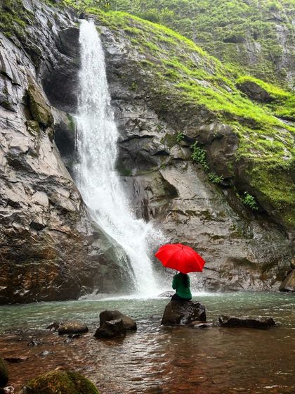 A beautiful shot of a trekker with a red umbrella sitting in front of a waterfall, announcing our upcoming monsoon trek in Maharashtra.
