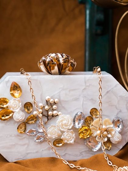 This white marble-effect acrylic clutch is adorned with golden-hued rhinestones and delicate 3D floral accents. It includes a detachable gold chain sling for versatile styling.