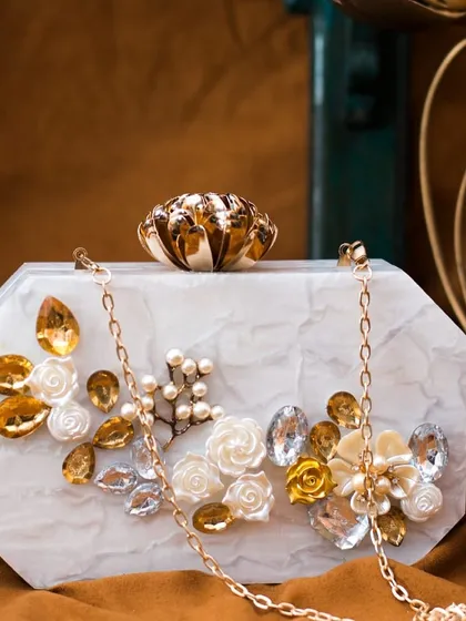 This white marble-effect acrylic clutch is adorned with golden-hued rhinestones and delicate 3D floral accents. It includes a detachable gold chain sling for versatile styling.