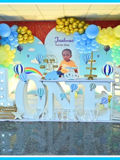 A dreamy hot air balloon-themed first birthday. The stage is set with a large 'ONE' marquee table, a teddy bear in a balloon-basket, and a sky-blue backdrop.