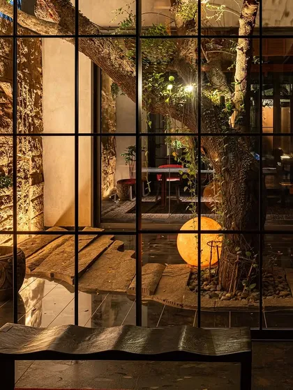 A view through a large window into the KEUS courtyard at night. The illuminated tree and glowing orb create a captivating, gallery-like scene from the inside.