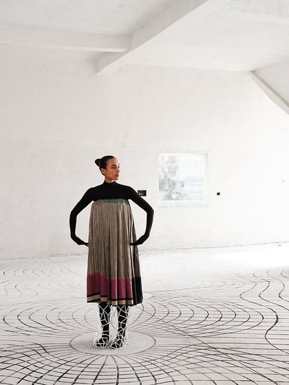 A lone figure stands in a vast, white room with a swirling floor pattern. The styling is simple to draw attention to the interaction between the body and the space.