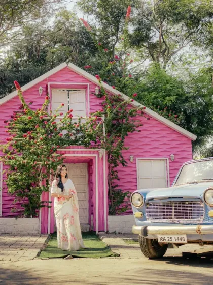 A wide shot that tells a story. The groom waits by the vintage car as his bride emerges from the charming pink house, setting the scene for their romantic, retro-themed pre-wedding shoot.