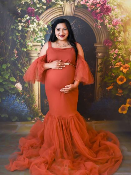 A beautiful studio portrait of a mom-to-be in a rust-colored gown, cradling her baby bump in front of a floral arch.